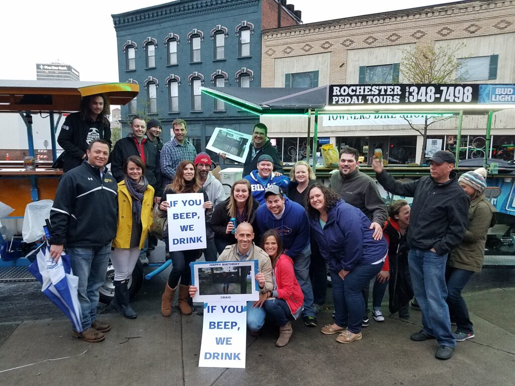 group of friends having fun posing for a picture in front of two pedal pubs in the city holding signs that are saying "If you beep, we drink"