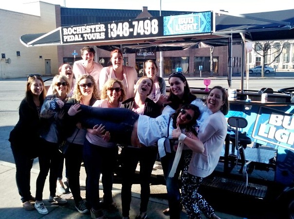 group of ladies holding their friend in the air horizontally posing for a picture in front of a pedal pub