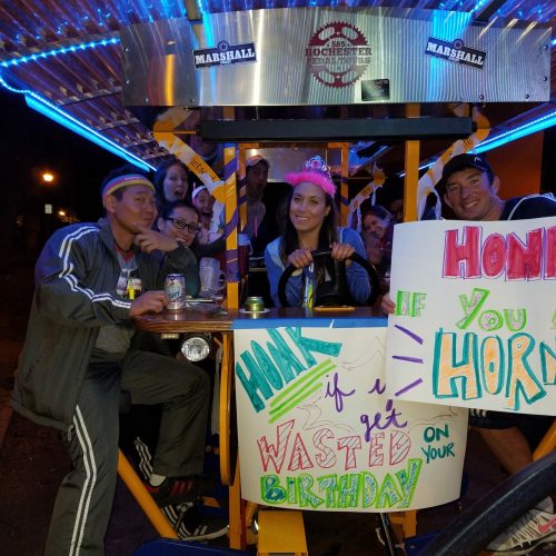 friends taking a picture while driving a pedal pub celebrating a birthday of a lady that is holding the wheel of the pedal pub