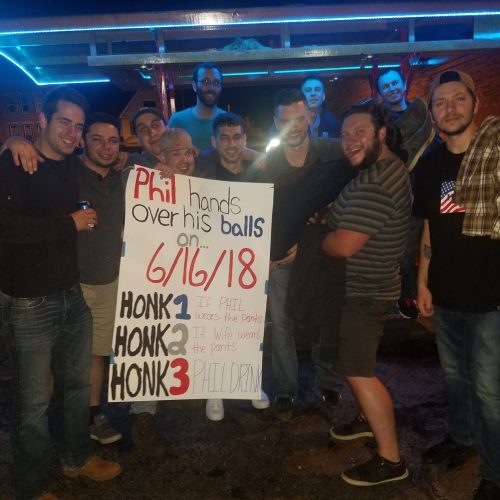 group of guys smiling and posing in fron of a pedal pub holding a sign celebrating their friends marriage