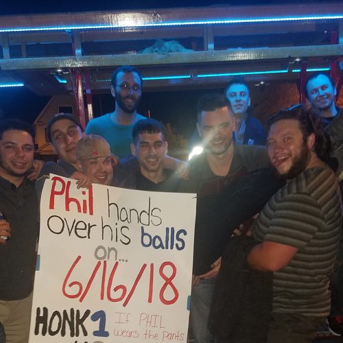 group of guys smiling and posing in fron of a pedal pub holding a sign celebrating their friends marriage