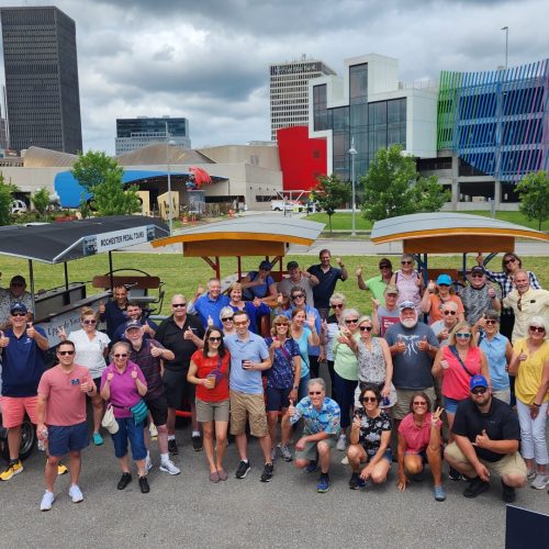 large group of people posing for a picture holding thumbs up in fron of 3 pedal pubs in the city parking lot